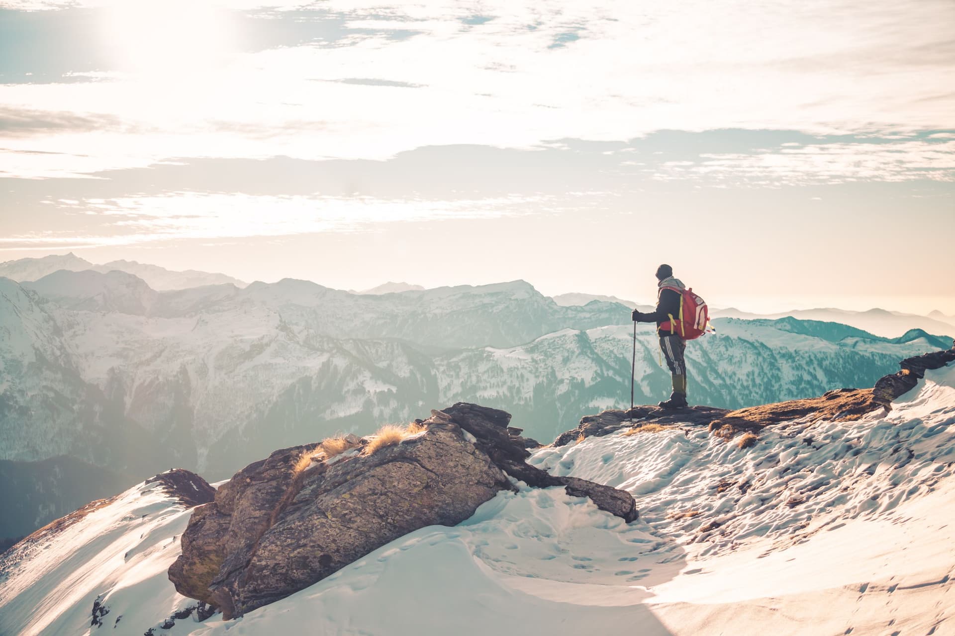 A lone trekker with a pole and pack on a snow ridge at sunrise, looking out over the ranges