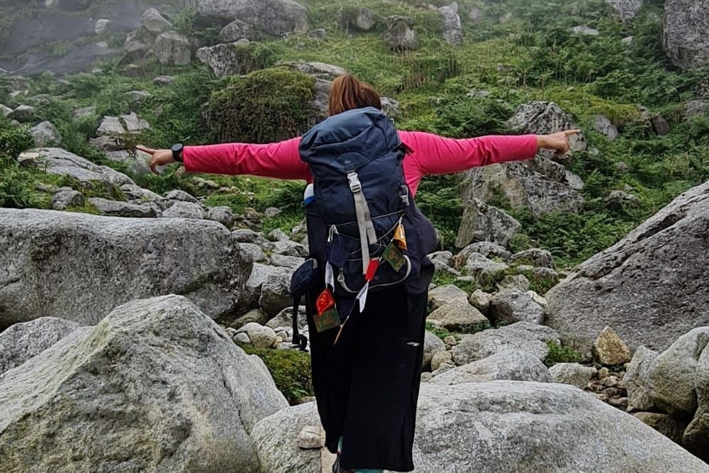 A trekker with a backpack spreading arms in joy while hiking through rocky terrain on the Kareri Lake Trek in Himachal Pradesh.