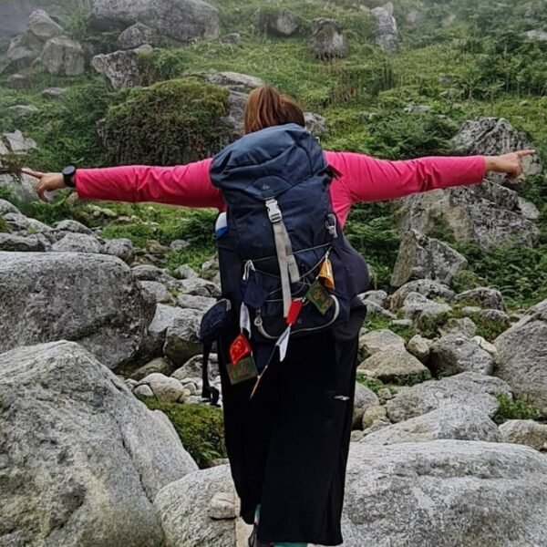 A trekker with a backpack spreading arms in joy while hiking through rocky terrain on the Kareri Lake Trek in Himachal Pradesh.